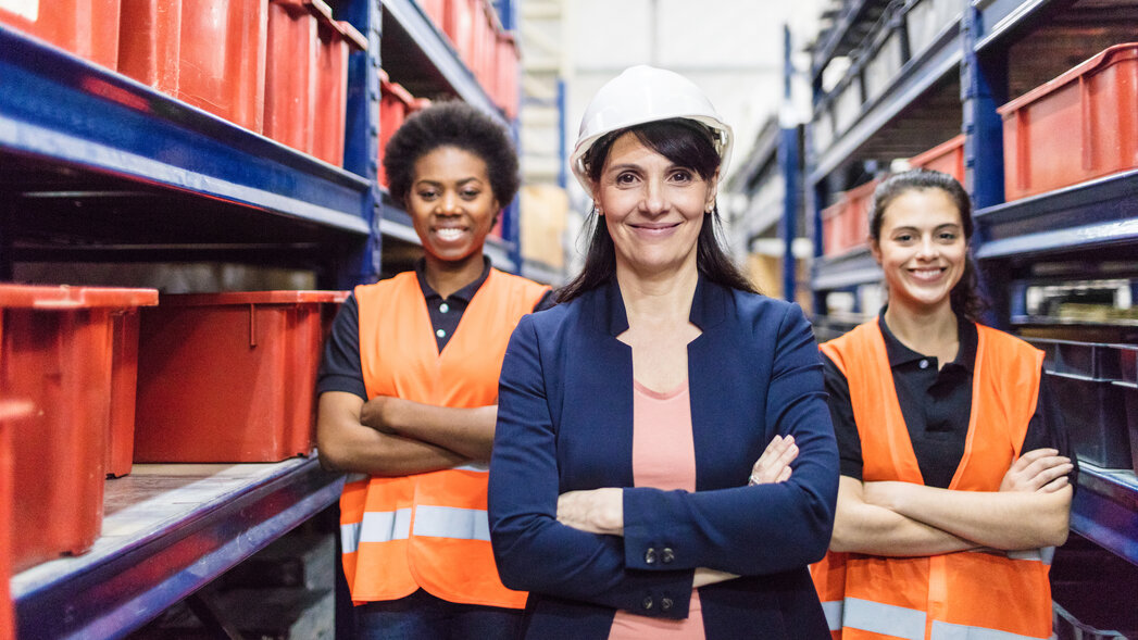 Drei selbstbewusst wirkende und lächelnde Frauen befinden sich in einem Lagerhaus. Eine trägt einen Schutzhelm.
